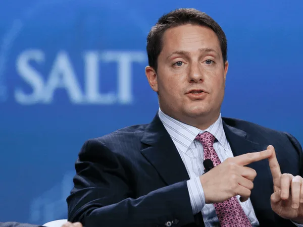 Male conference speaker in a suit, gesturing with his hands on a blue backdrop with a partial 'SALT' logo behind him
