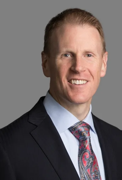 Professional headshot of a smiling man in a dark suit, light blue shirt, and paisley tie against a gray background.