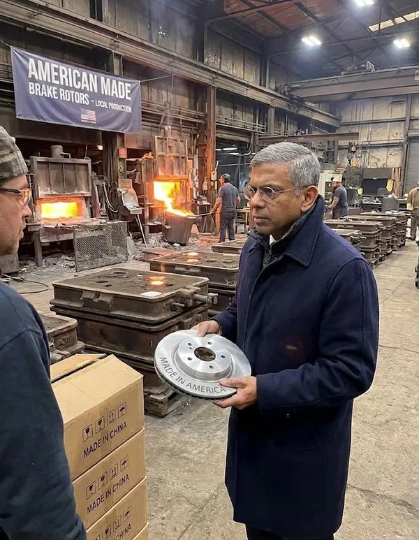 Man in a dark coat inspecting a silver brake rotor stamped 'Made in America' in a metal forging shop with molten furnaces in the background.