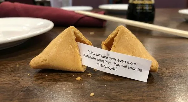 Two fortune cookies on a wooden table; a fortune reads: 'China will take over even more American industries. You will soon be unemployed.'