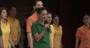 Cecilia Salinas [daughter of Carlos Salinas] addresses her cult leader Keith Raniere at his birthday. She is wearing a green tee shirt because it signifies her high rank in Nxivmn [Green Sash]. Others are wearing orange or yellow based on their rank.