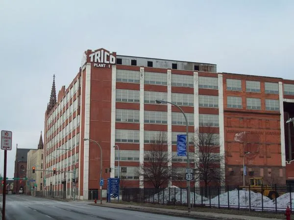 Red brick industrial building with white columns and large TRICO Plant 1 sign on the top corner, street in front with traffic lights and a fence.