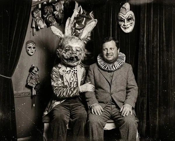 Two men seated on a bench in a theater setting wearing eerie costumes: one with a worn rabbit head mask, the other in a suit with a ruffled clown collar, surrounded by hanging masks and puppets.