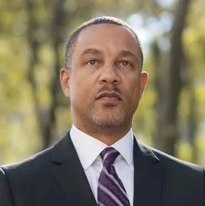 Head-and-shoulders portrait of a man in a dark suit and purple striped tie outdoors, with blurred trees in the background.